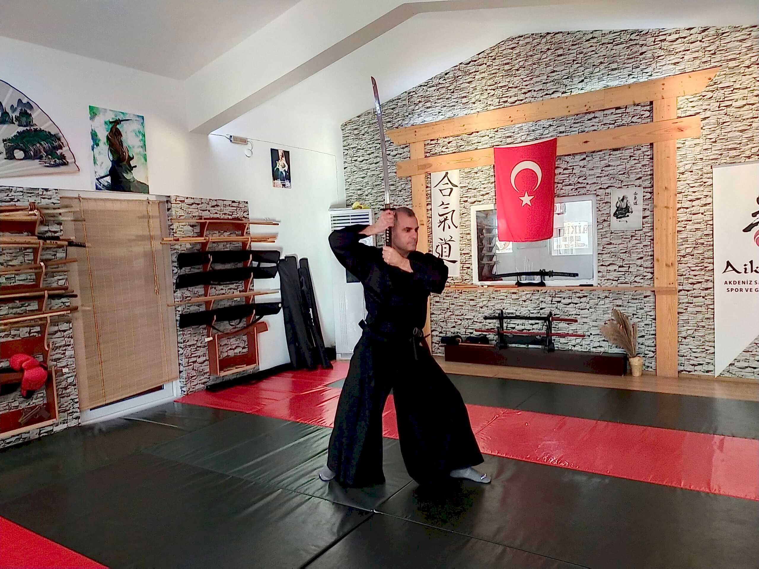 Kürşat Demir, geleneksel siyah kıyafetiyle, Türkiye bayrağı ve aikido sembolleri asılı bir dojo'da katanasını kınından çıkarıyor.
